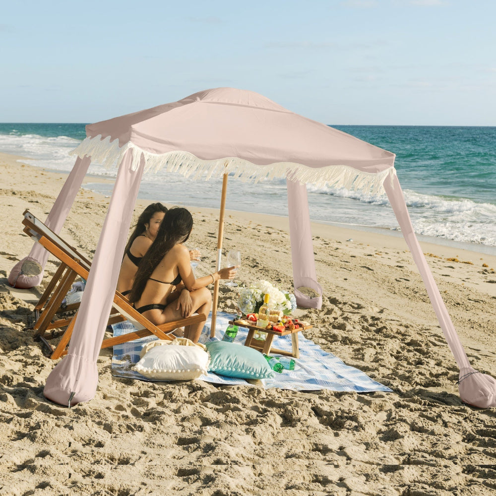 Two women sitting in the AMMSUN 7'×7' Boho Beach cool cabana beach tent with Fringe,Pink Coral canopy