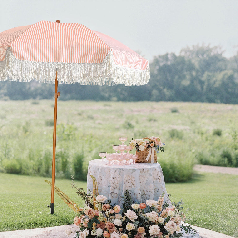 Outdoor setting with a table decorated with a cake, flowers, and a pink striped umbrella in a field.