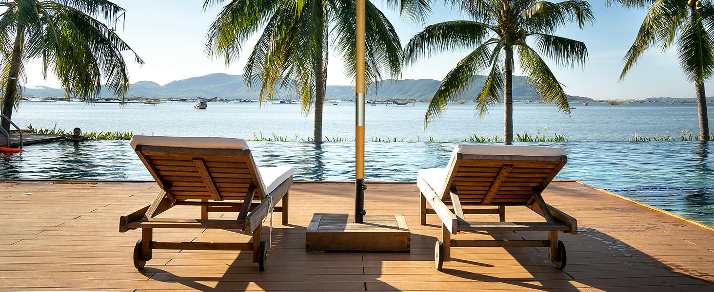 Two wooden lounge chairs on a wooden deck by a waterfront with palm trees and mountains in the background.