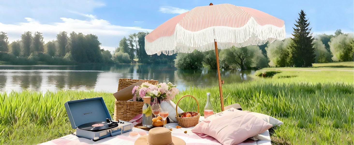 Picnic setup with a pink fringe umbrella outdoor patio, food, and a record player by a lake.