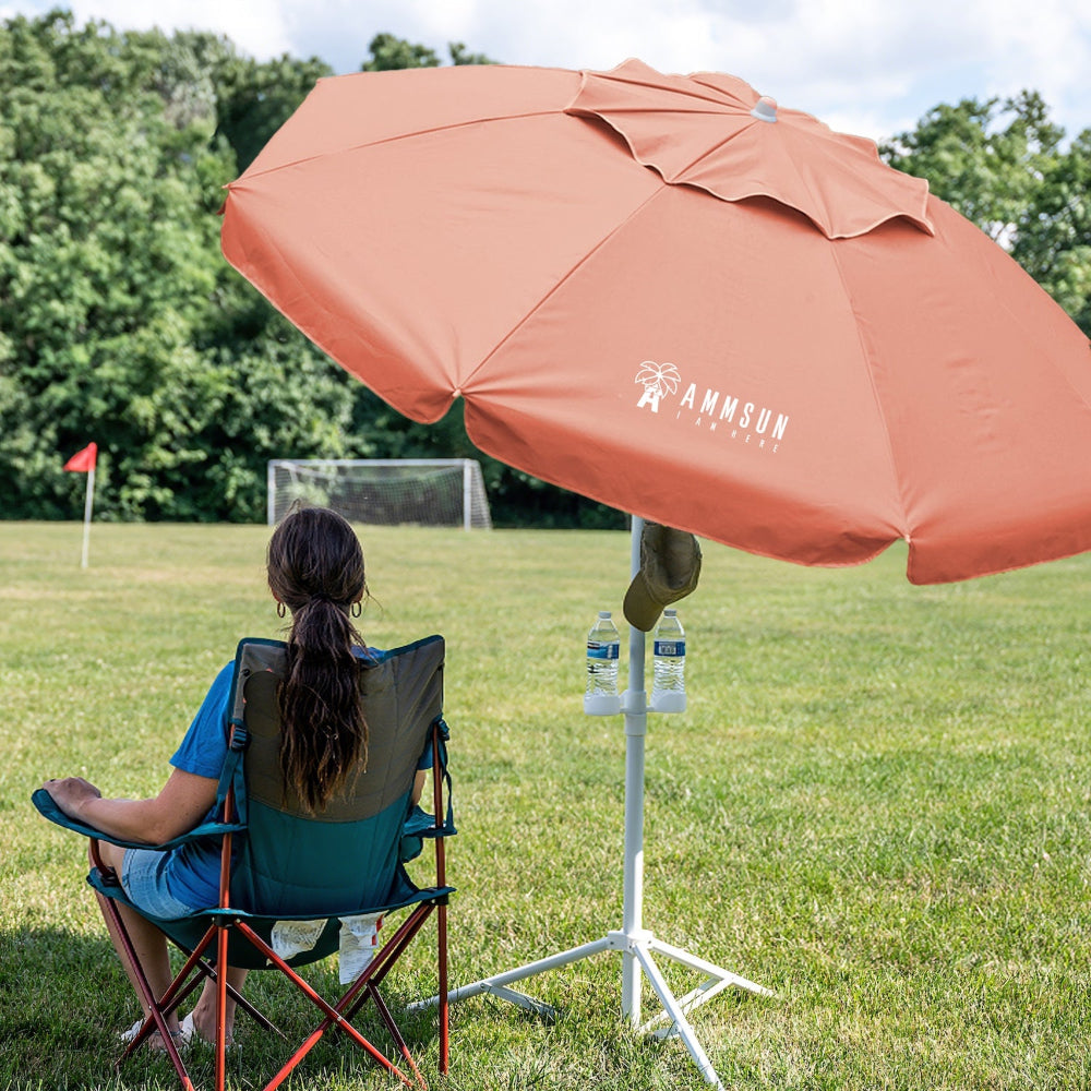 A woman sitting under a large orange sports umbrella with stand for outdoor umbrellas on grass.