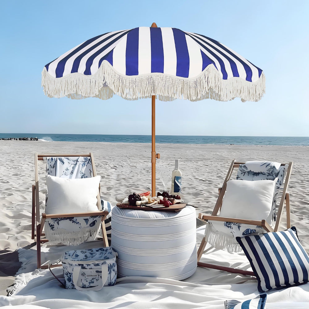 Beach setting with striped portable beach umbrella with fringe, chairs, and a table with snacks on a sandy beach.
